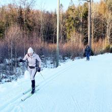 Dame som går på ski i skogen med mann bak.